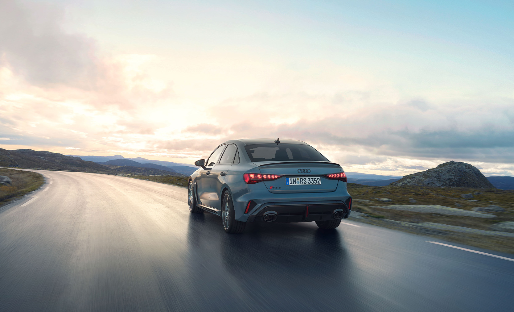Die grauer Audi RS3 Limousine in der Heckansicht, wegfahrend auf einer nassen Stra&szlig;e und H&uuml;gellandschaft.