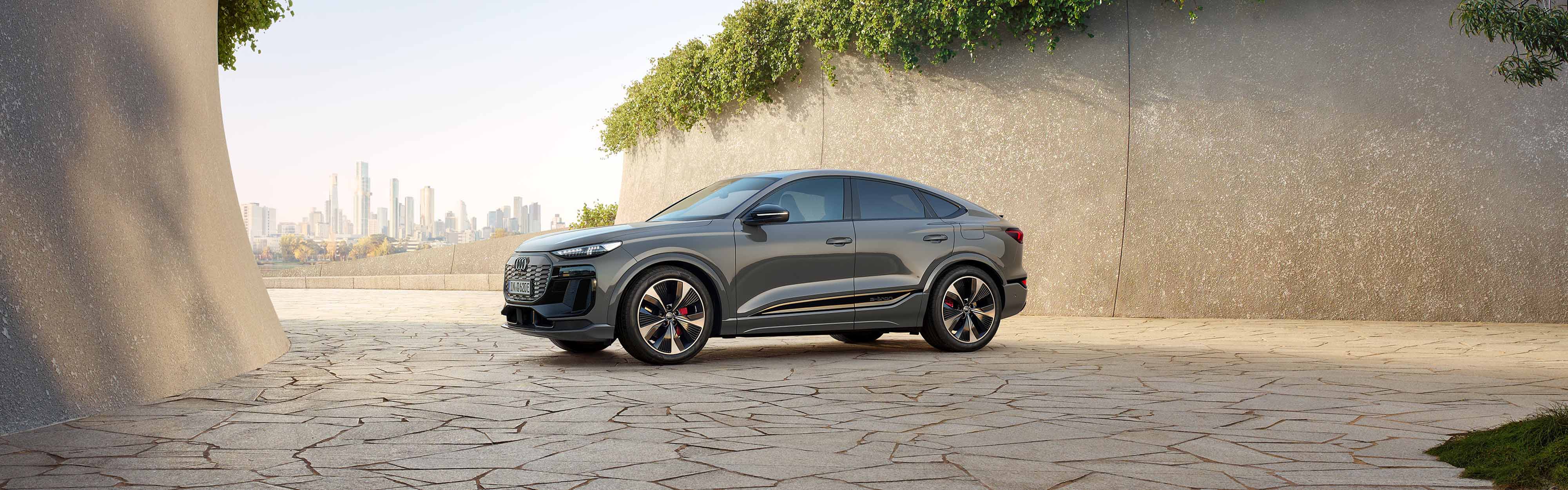 Der neue Audi Q6 Sportback e-tron in der Seitenansicht zwischen zwei Steinigen Bergen. Im Hintergrund eine Stadt