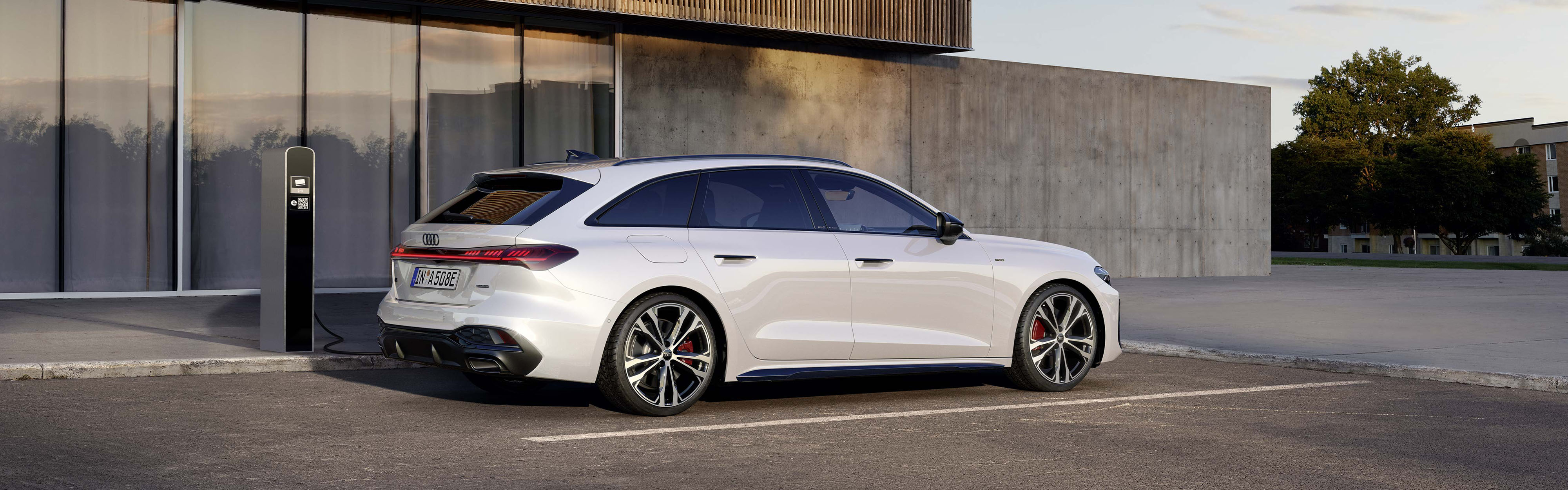 ein wei&szlig;er Audi A5 Avant e-hybrid an einer Lades&auml;ule vor einem futuristischem Geb&auml;ude