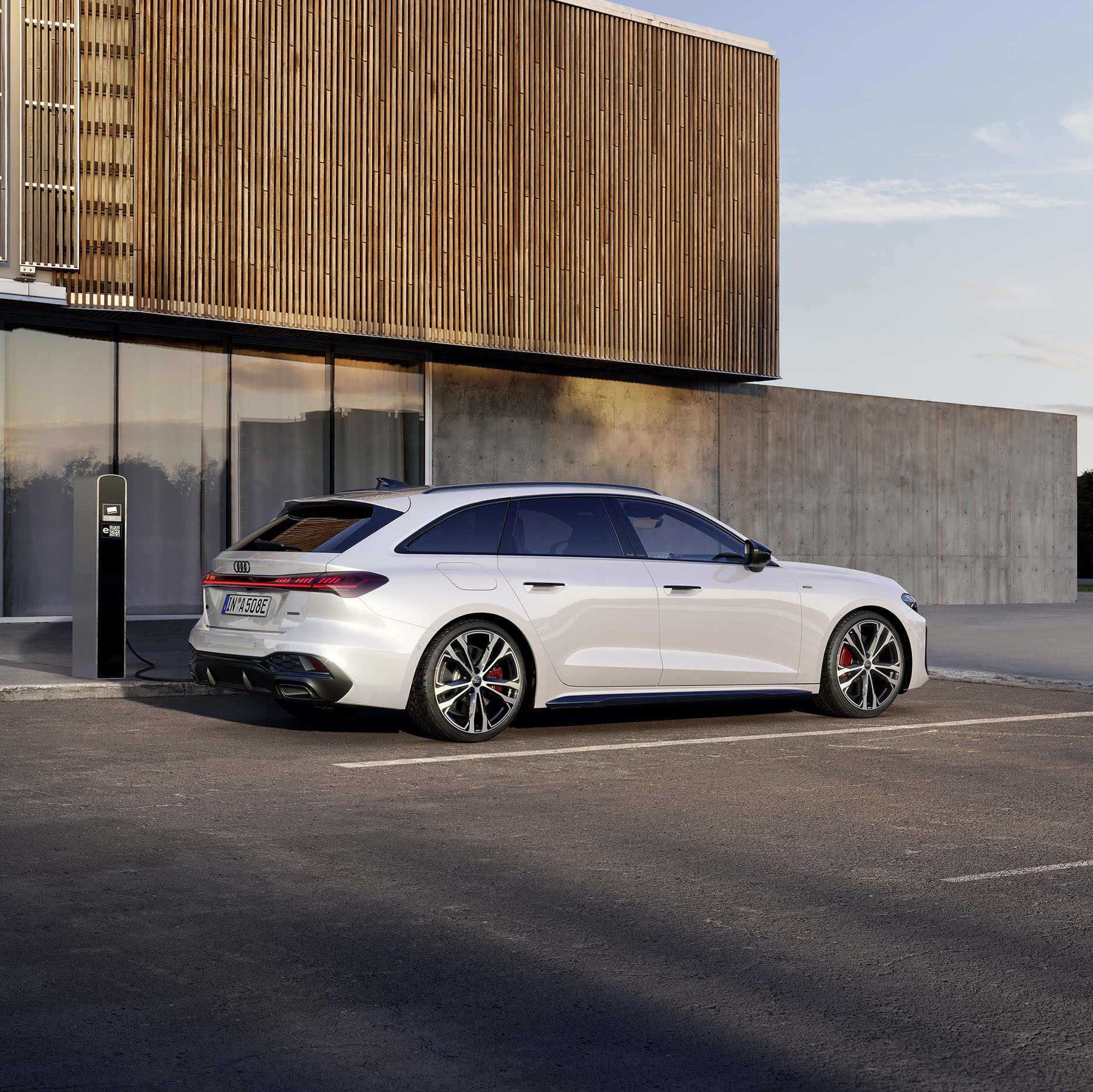 ein wei&szlig;er Audi A5 Avant e-hybrid an einer Lades&auml;ule vor einem futuristischem Geb&auml;ude