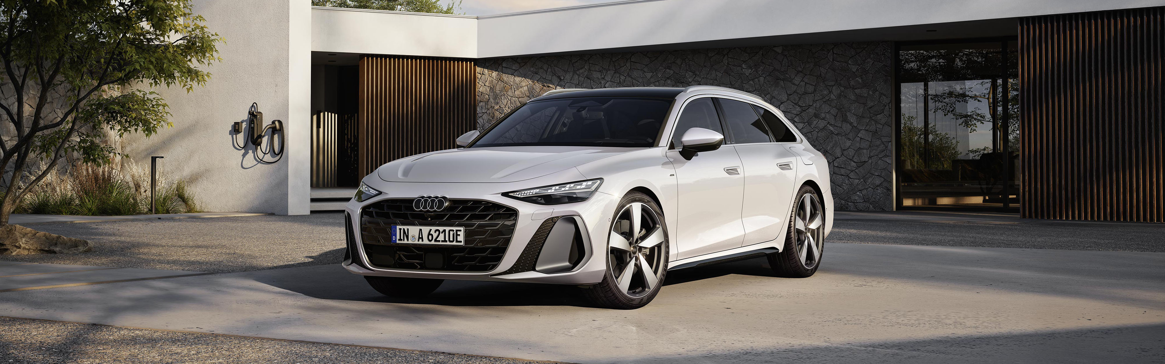 Ein wei&szlig;er Audi A6 Avant e-hybrid in der Dreiviertelansicht von vorn, vor einem futuristischem Geb&auml;ude