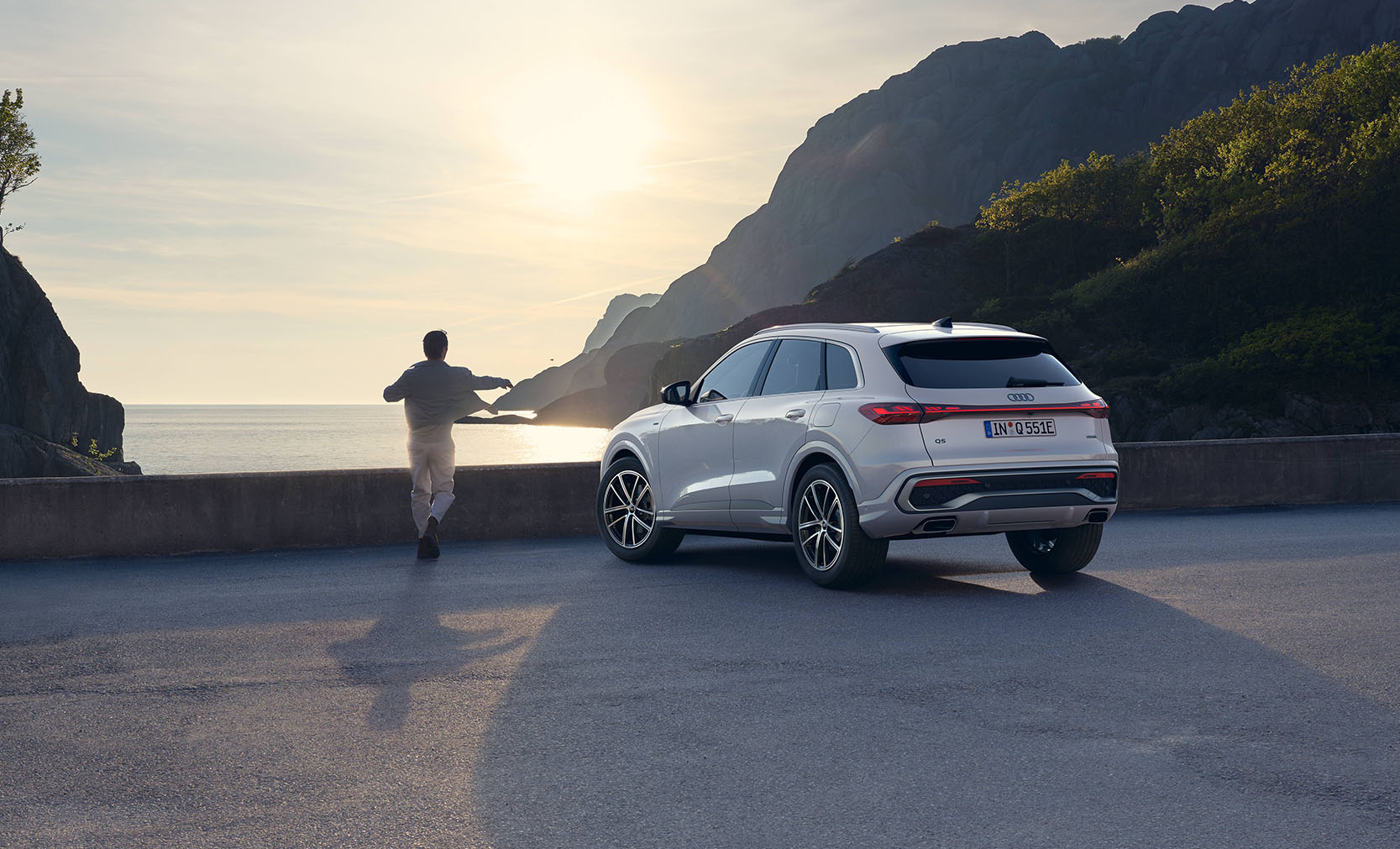 Ein wei&szlig;er Audi Q5 SUV e-hybrid von hinten mit einer Person vor einem K&uuml;stenabschnitt