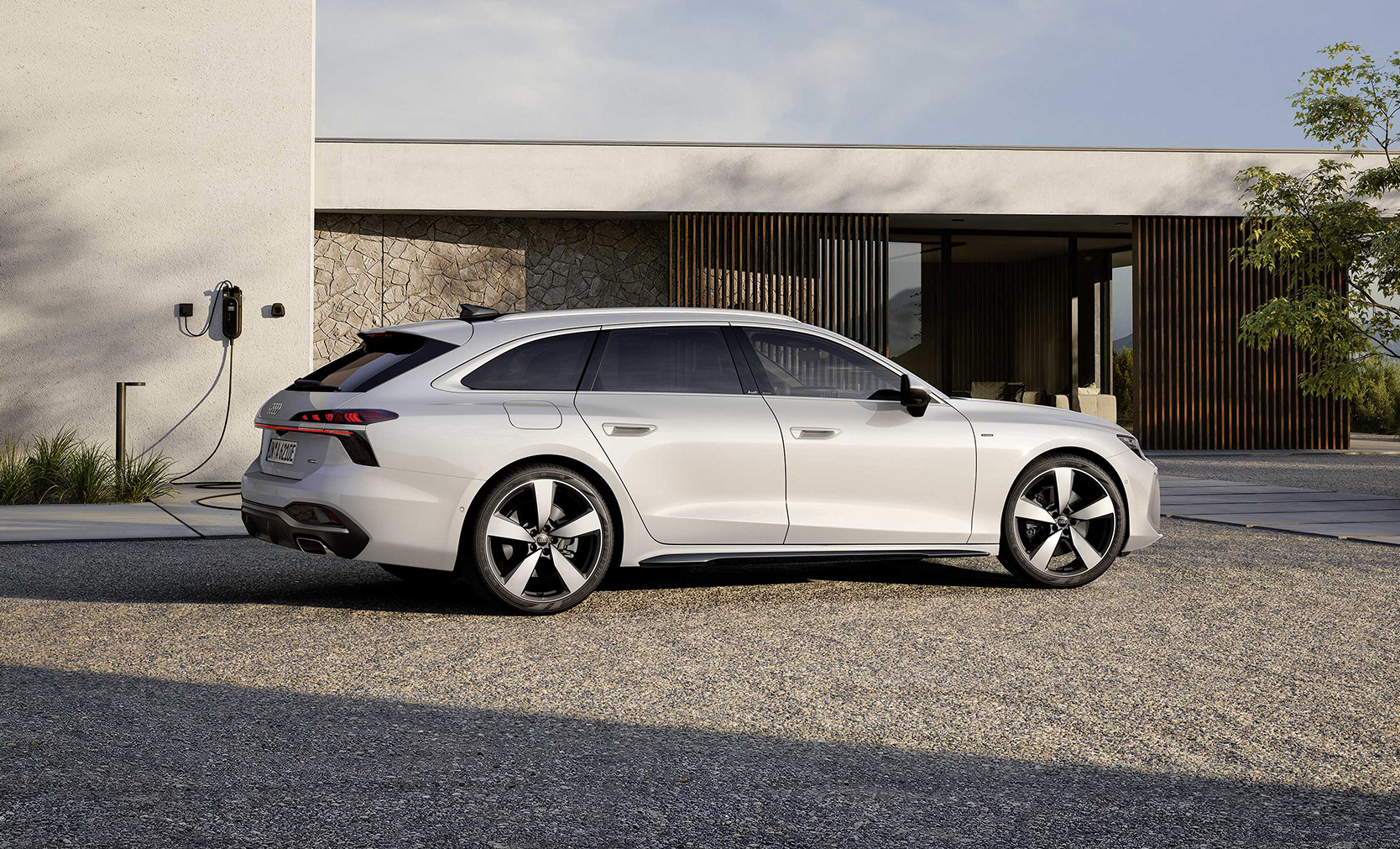 Ein wei&szlig;er Audi A6 Avant e-hybrid in der Dreiviertelansicht von vorn, fahrend einem futuristischem Geb&auml;ude